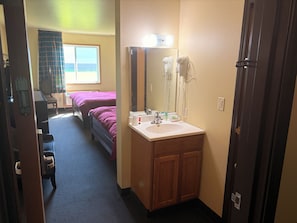 A hotel room with a bed, a desk, a chair, a bathroom with a sink and mirror, and a door.