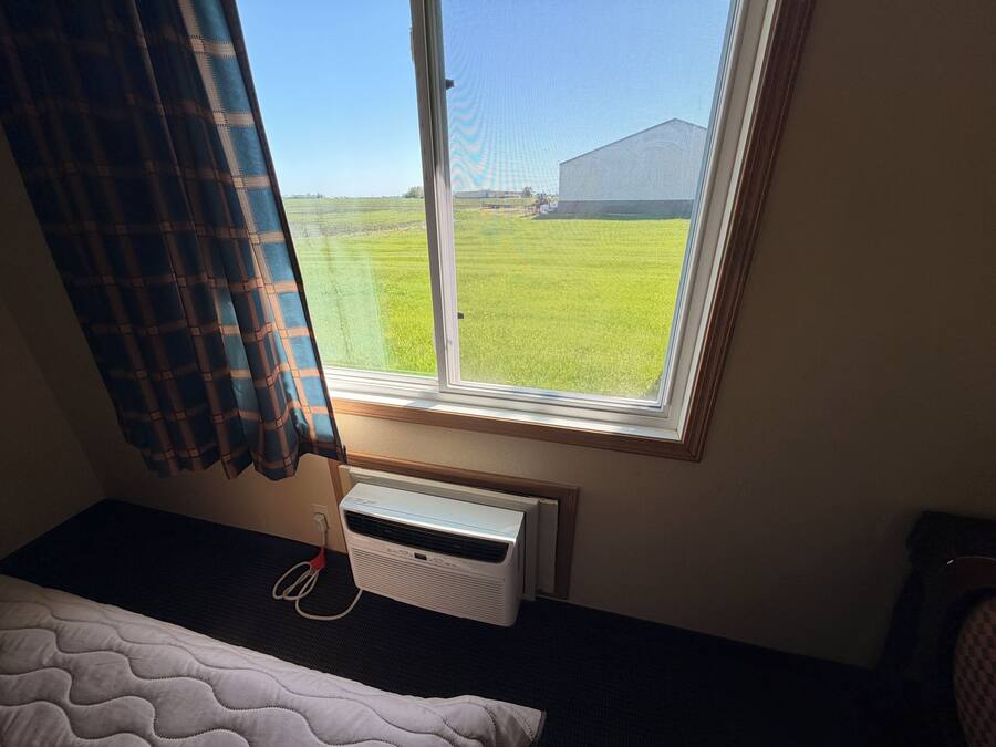 Una habitación con cama, ventana con cortinas, radiador y vistas a un campo y un edificio.