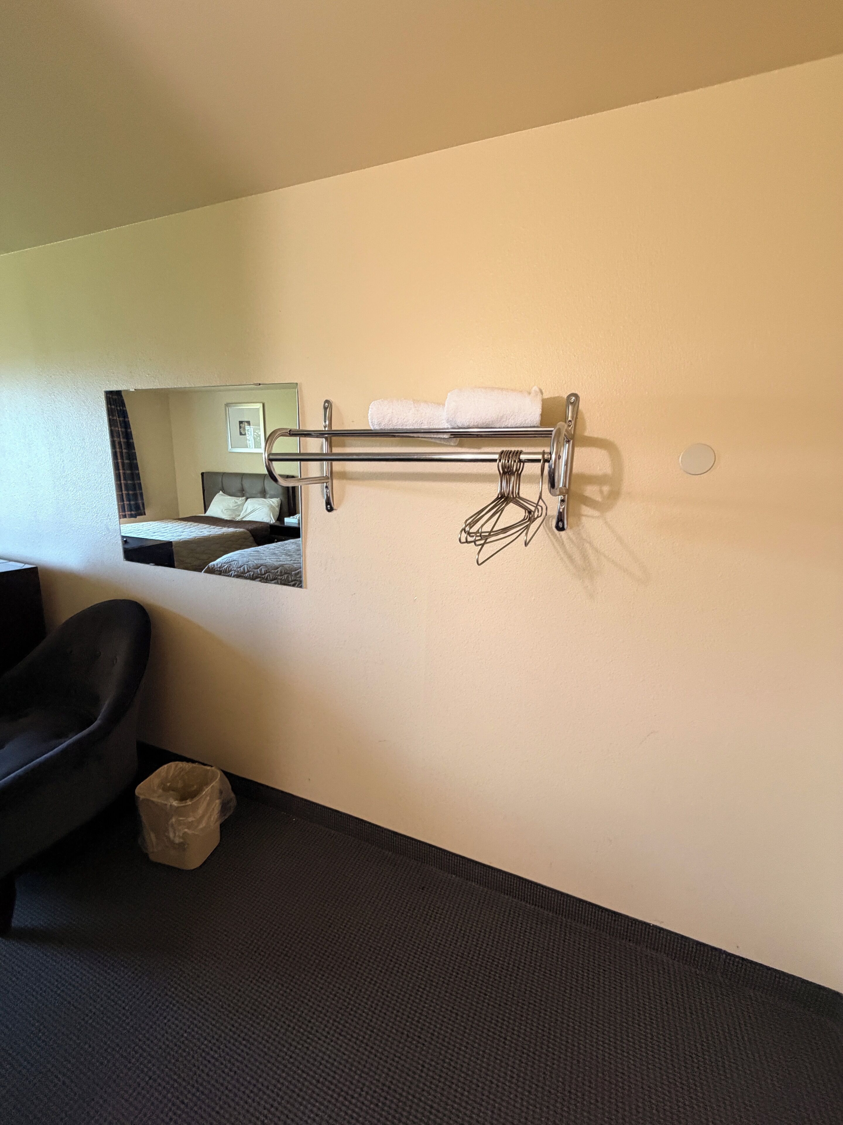 A hotel room with a bed, a black chair, a towel rack, and a small trash bin.