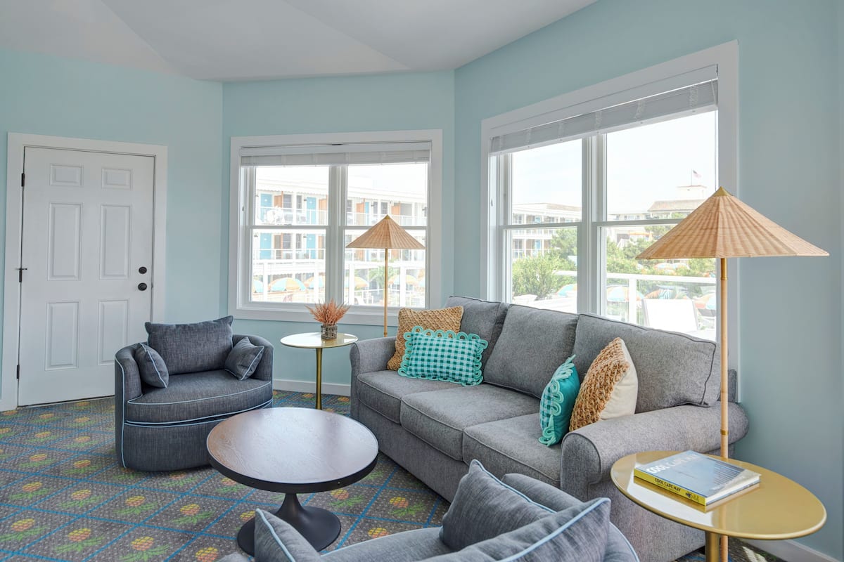 A living room with a sofa, armchair, and coffee table, large windows with blinds, and a view of buildings outside.