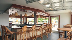 Restaurante com cadeiras de vime, área de bar e janelas amplas com vista para uma área externa ensolarada.