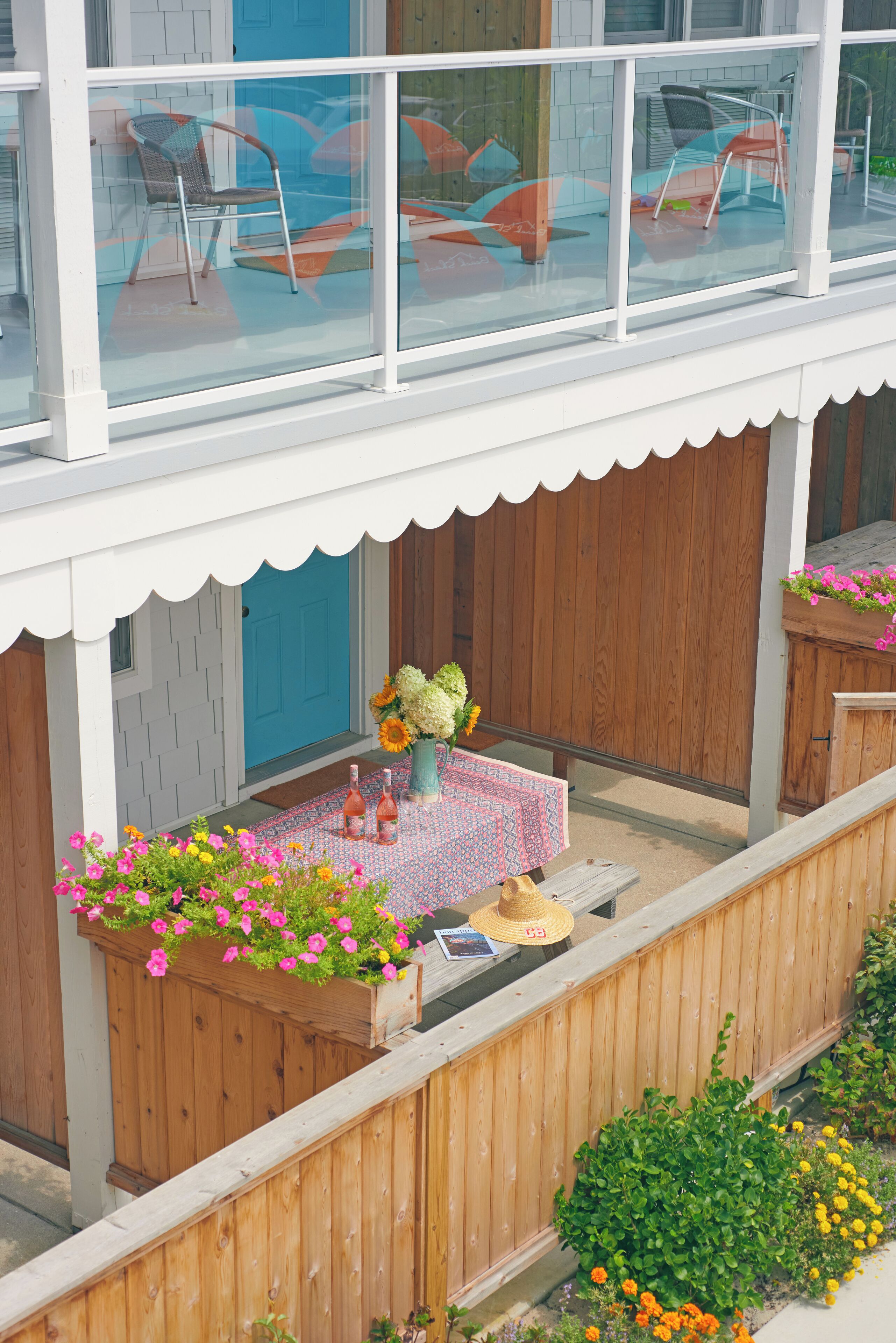 A covered patio with a table set for two, surrounded by wooden planters with flowers, a wooden fence, and a blue door.