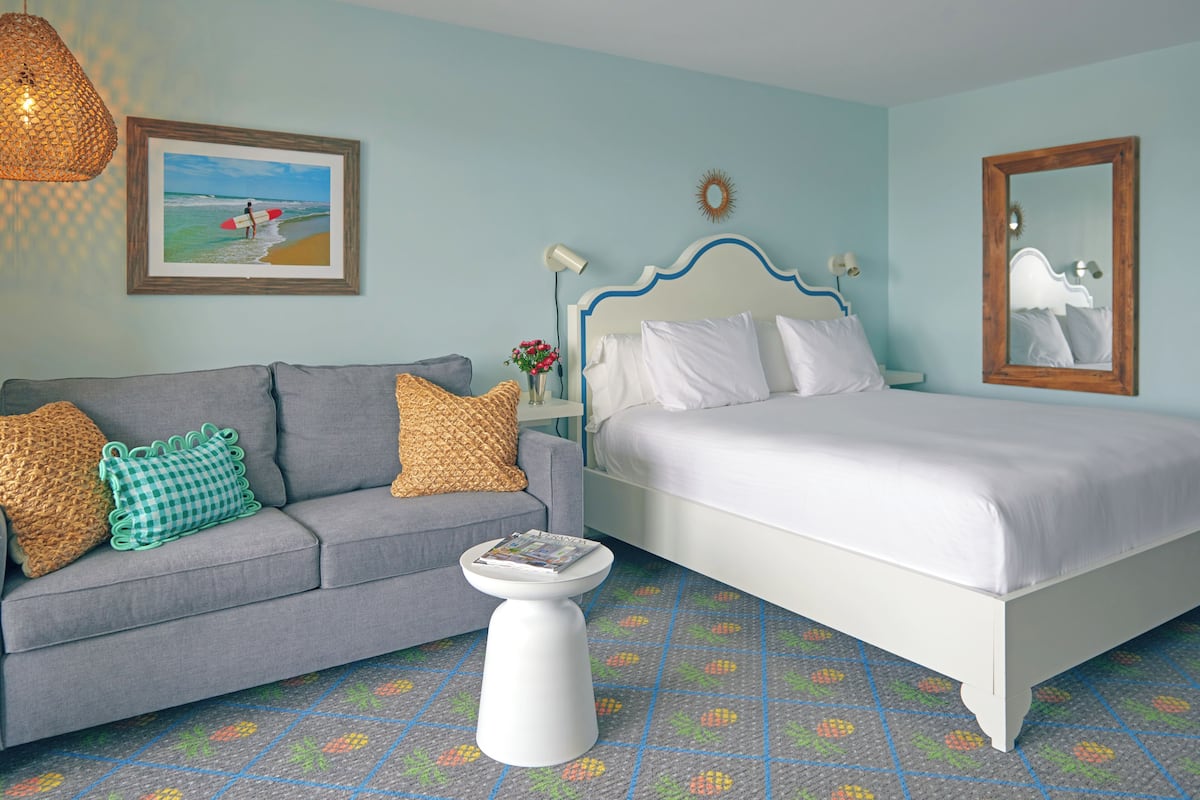 A bedroom with a bed, a sofa, a small table, a wall-mounted mirror, and a framed picture of a beach scene.