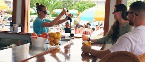 Un espace bar avec un comptoir, un panier de fruits et une vue sur une piscine agrémentée de parasols.
