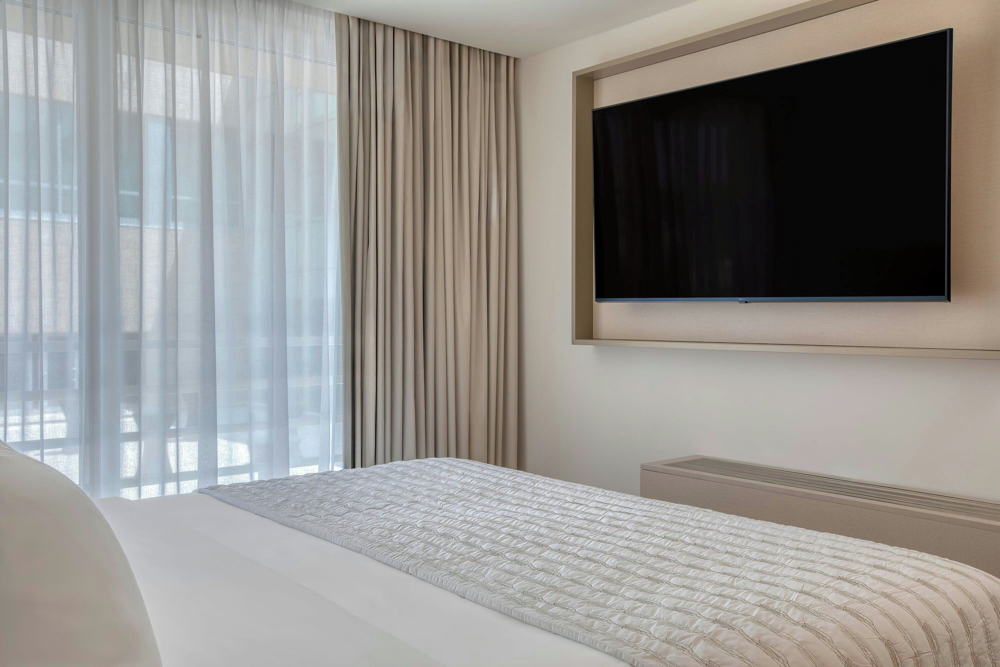 Habitación de hotel con una cama grande, un televisor de pantalla plana en la pared y cortinas translúcidas en la ventana.