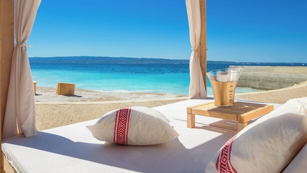 Una habitación frente a la playa con vista al mar, una mesa de madera con una bebida y una cama con almohadas.