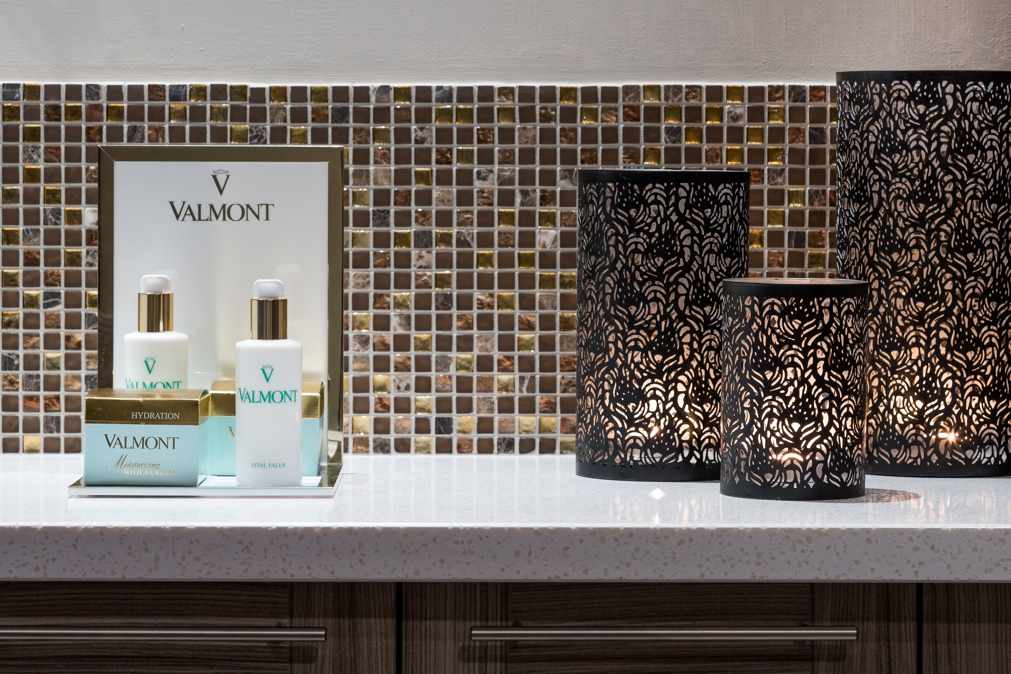 A bathroom vanity with a tiled backsplash, a framed Valmont skincare display, and decorative black candle holders.