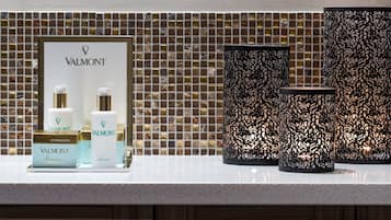 Un meuble-lavabo de salle de bain avec un pare-éclaboussures carrelé, un présentoir Valmont pour produits de soins de la peau, et des porte-bougies décoratifs noirs.