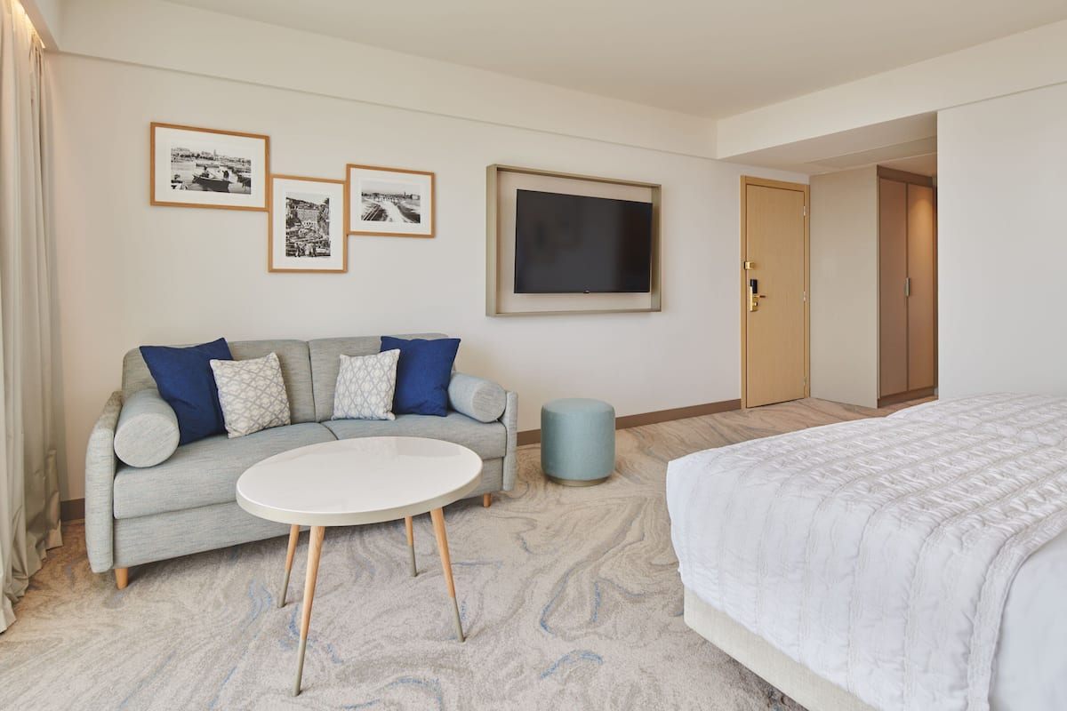 A hotel room with a grey sofa, a round white table, a bed with a white duvet, and framed pictures on the wall.