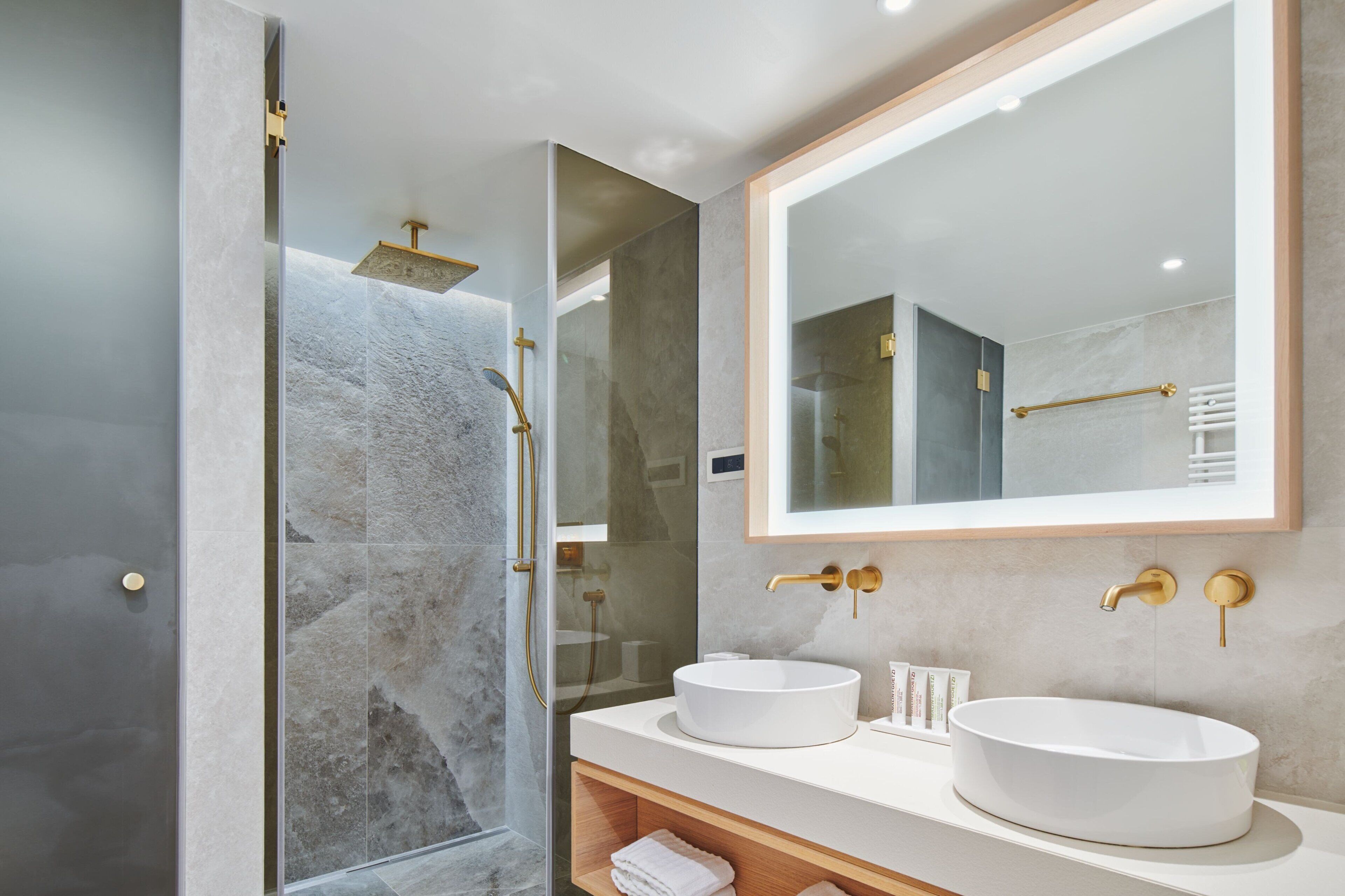 A modern bathroom with a glass-enclosed shower, two white sinks, and a large mirror with gold fixtures.
