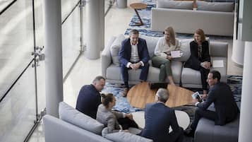 Un groupe de personnes est assis sur un canapé moderne de couleur claire, dans un hall spacieux et bien éclairé, doté d’un sol en marbre et d’un tapis à motifs.