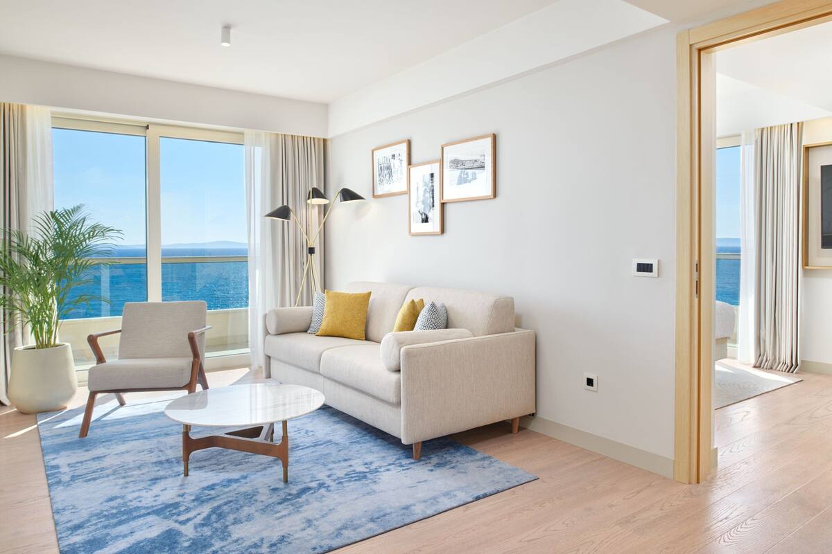 A modern living room with a sofa, armchairs, a coffee table, and a large window offering a view of the sea.