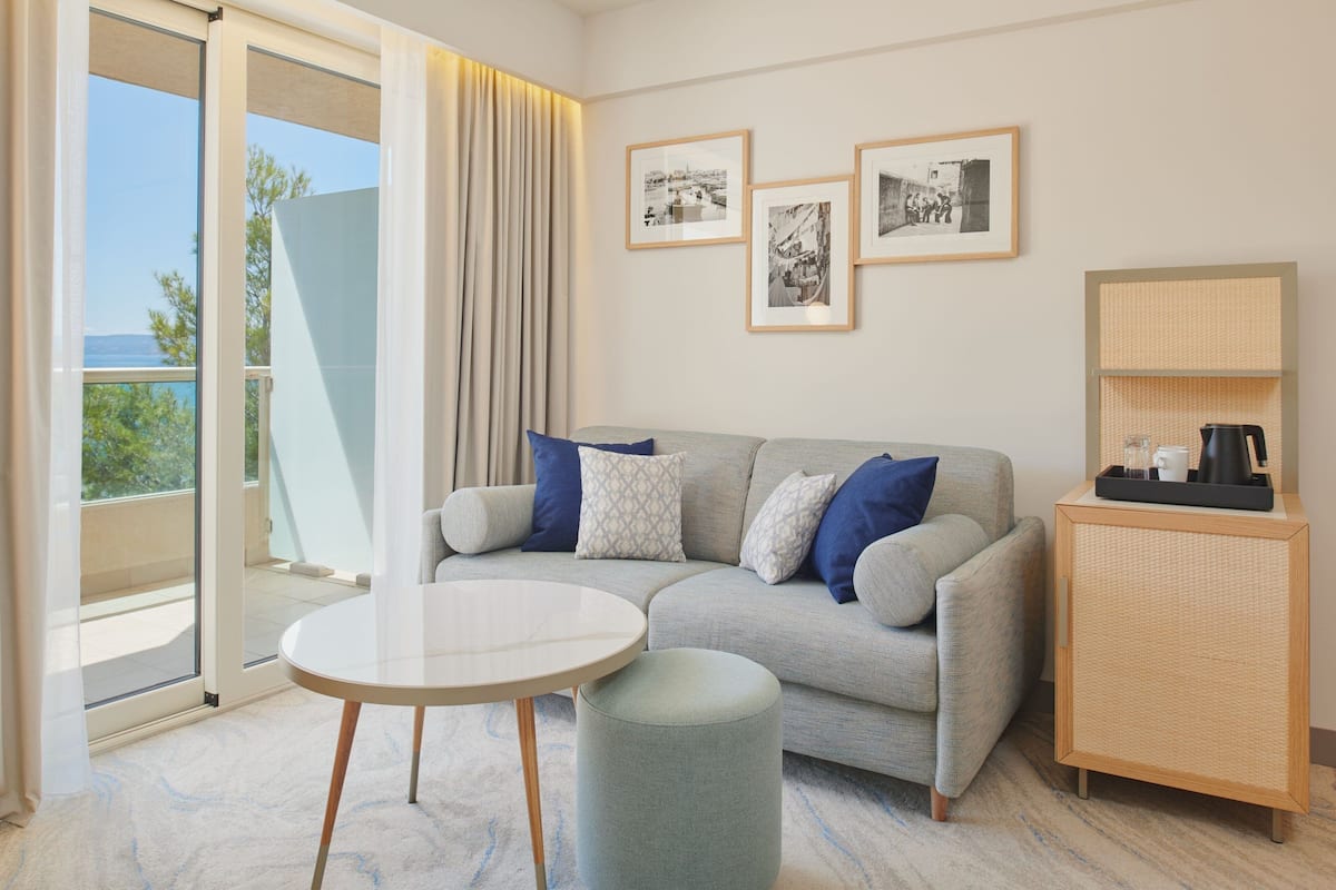 A modern hotel room with a grey sofa, a round table, and a small cabinet. There are three framed pictures on the wall and a view of the outdoors through the sliding glass door.