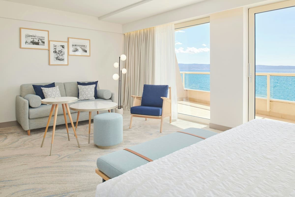 A hotel room with a bed, a sofa, a chair, a small table, and a view of the sea.