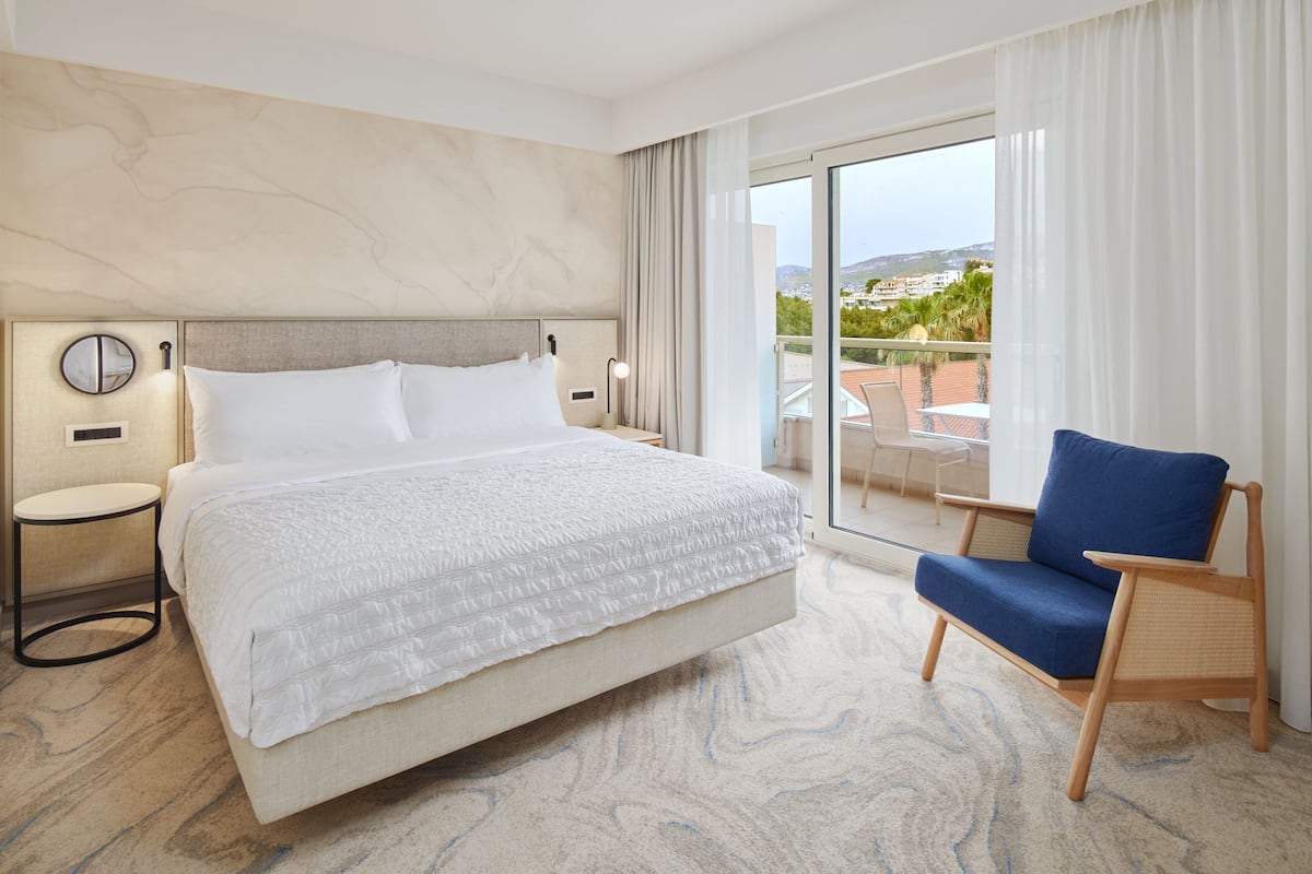 A hotel room with a large bed, a chair, a round table, and a view of the outdoors.