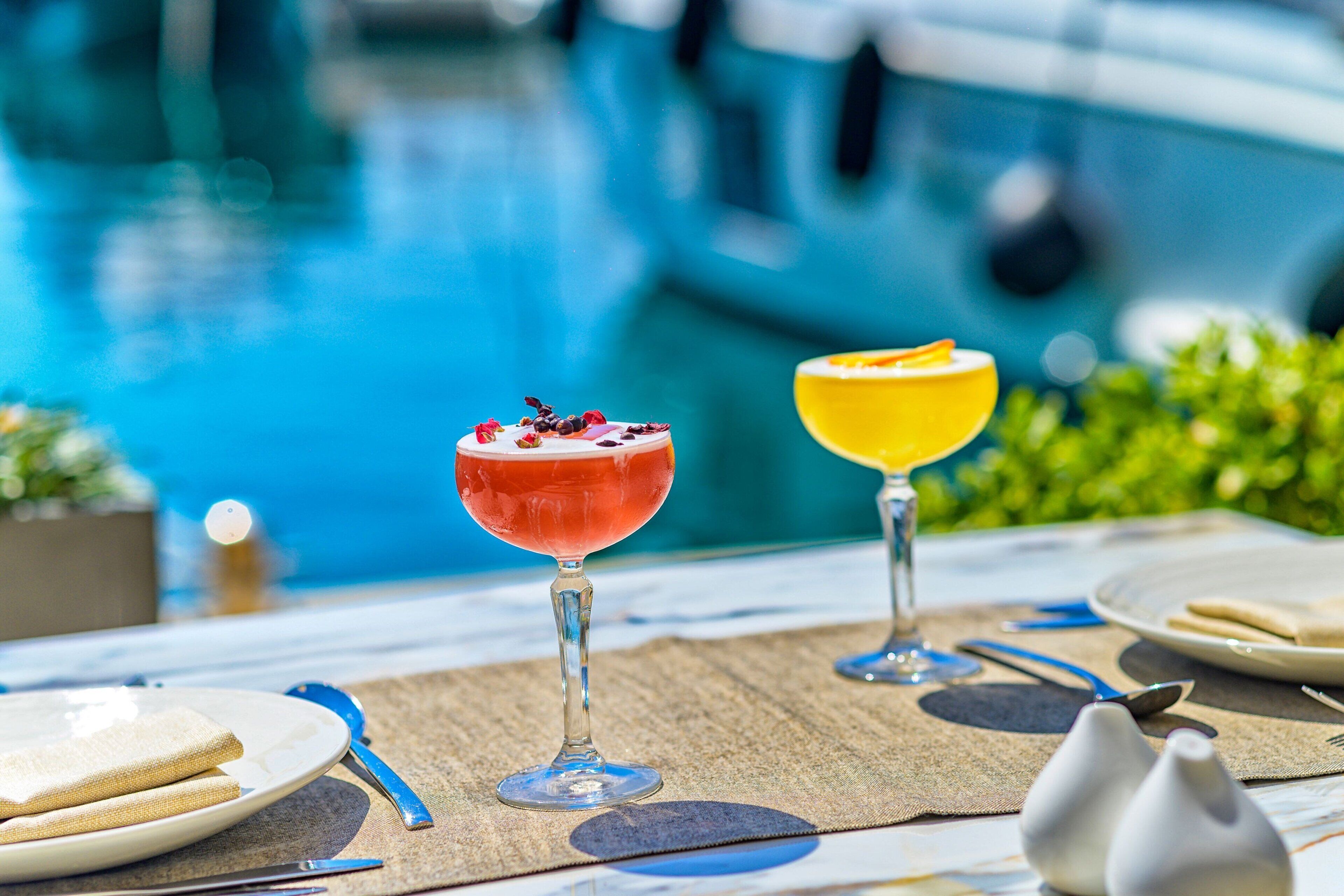 Una mesa con dos bebidas, un plato de sándwiches y una piscina al fondo.