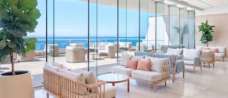 A modern lounge area with a view of the ocean, featuring sofas, chairs, and a large potted plant.
