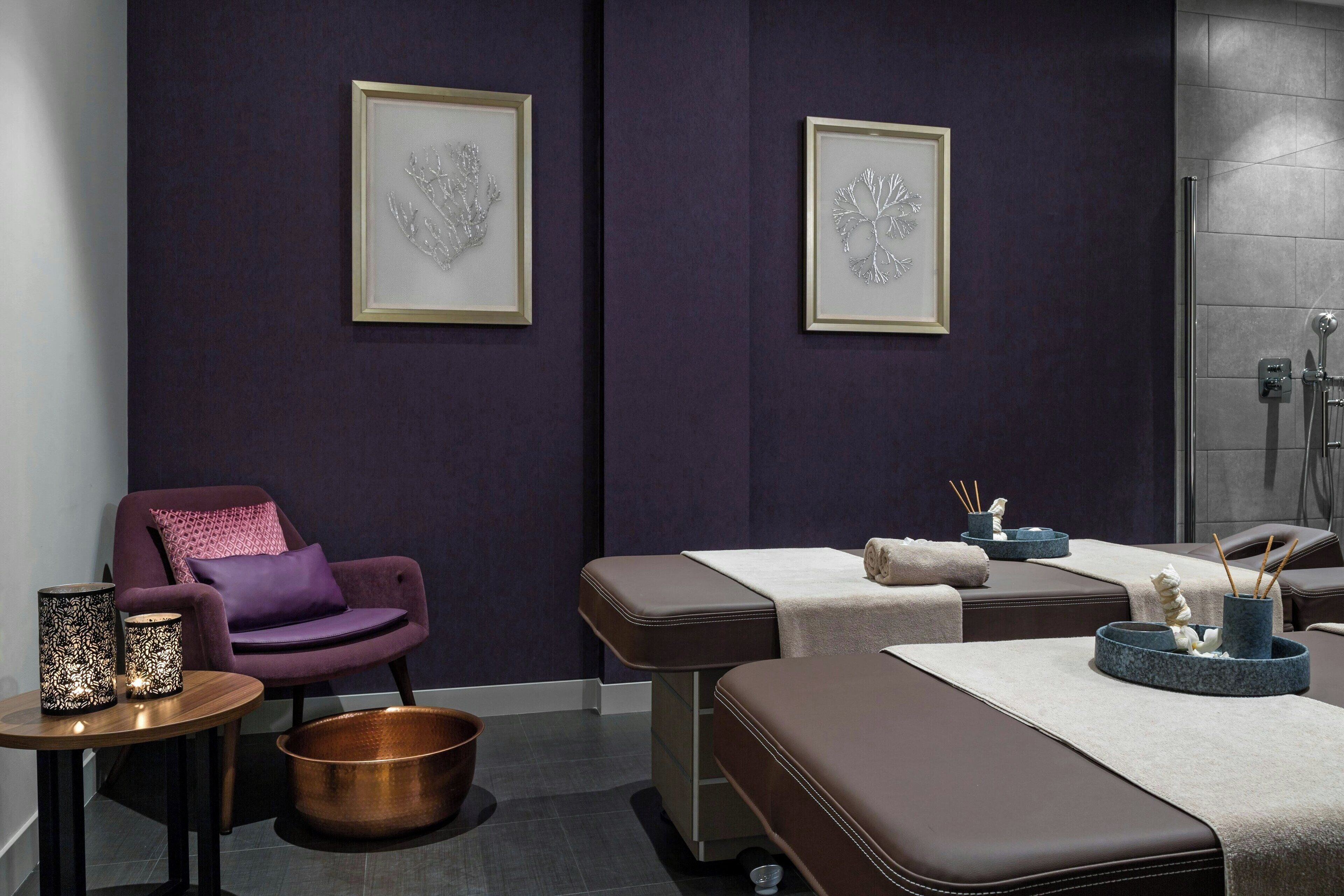 Een spa-ruimte met een paarse fauteuil, een massagetafel, kunstwerken in lijsten en een koperen emmer.