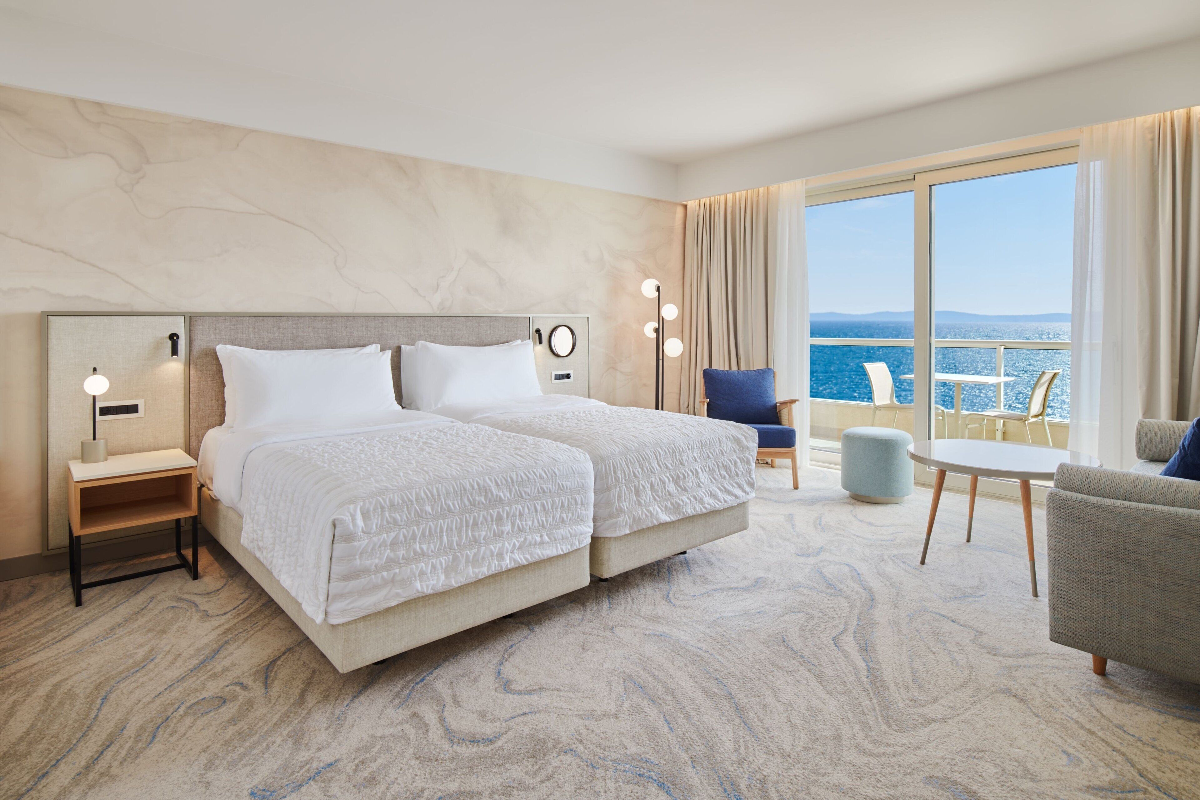 A hotel room with a large bed, a desk, a chair, and a balcony with a view of the sea.