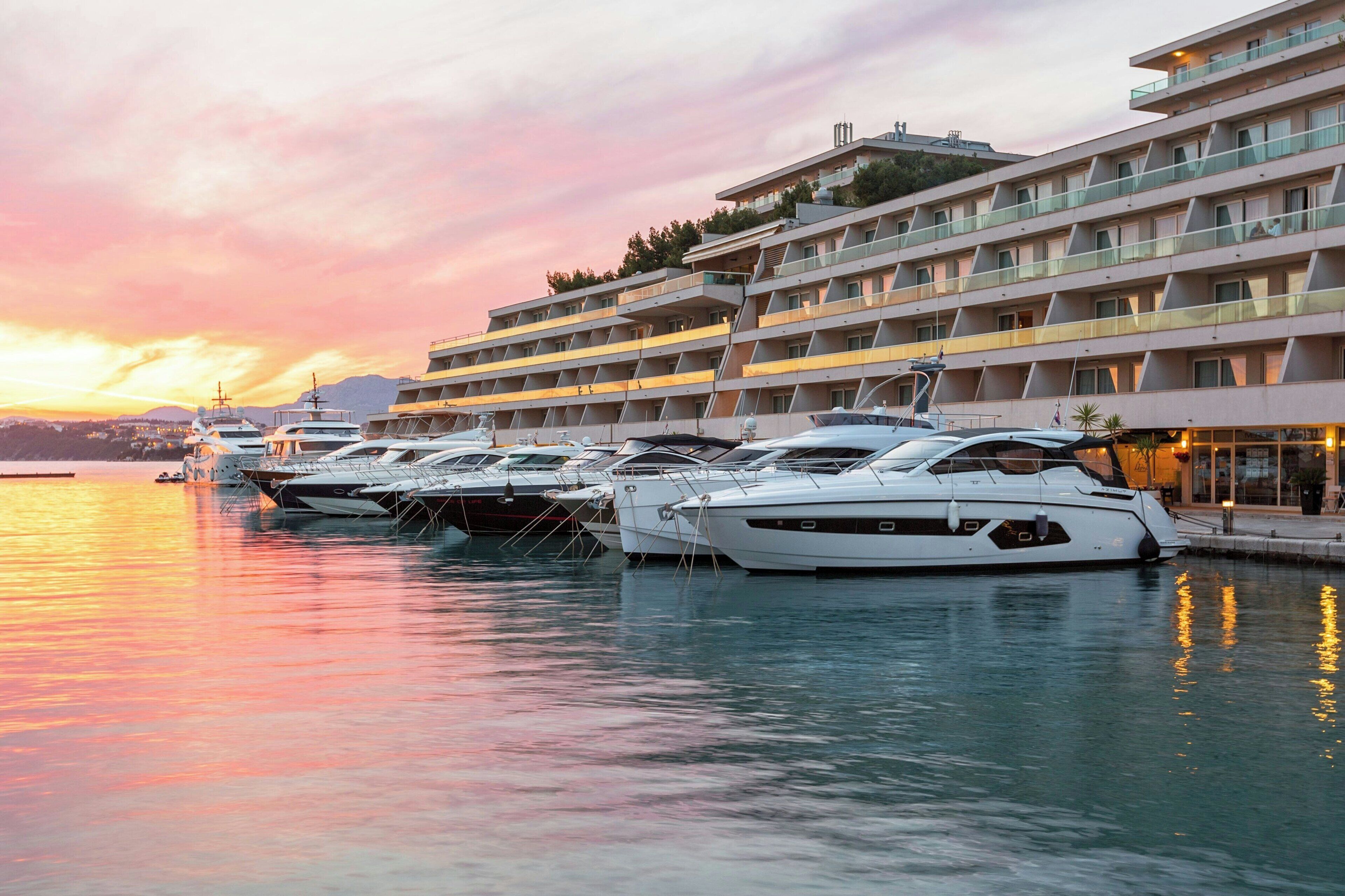 A marina with several yachts docked at a hotel with multiple balconies and a view of the sunset.