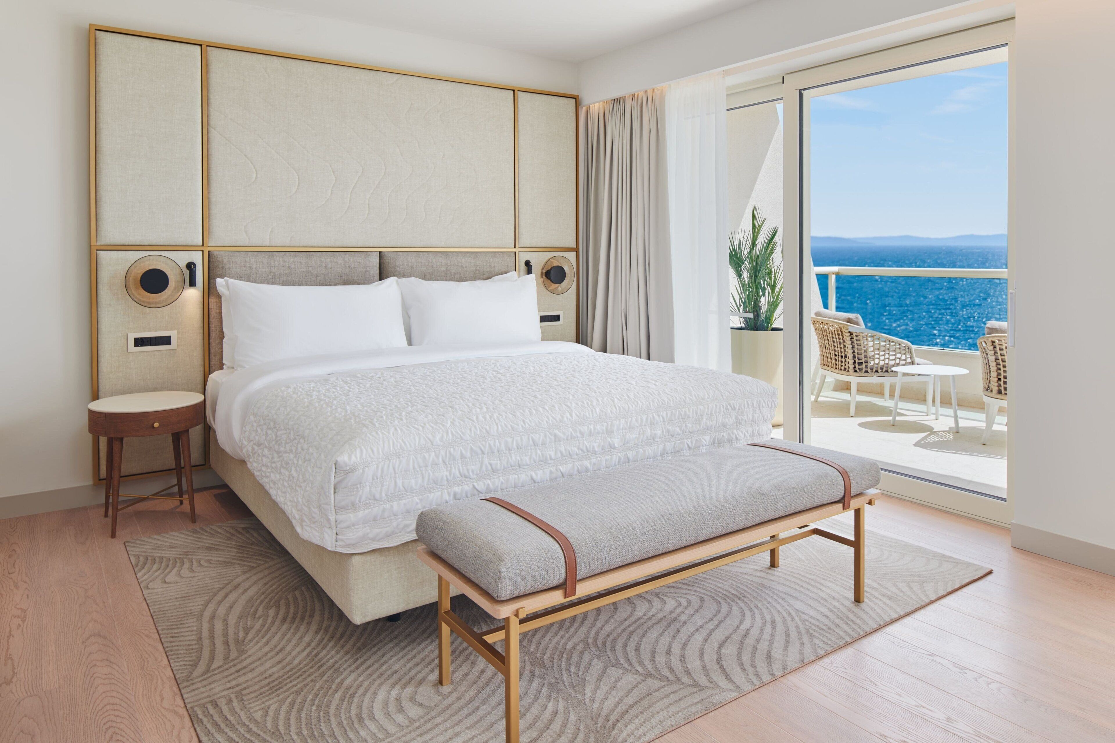 A hotel room with a large bed, a bench, a small table, and a view of the sea through a balcony.