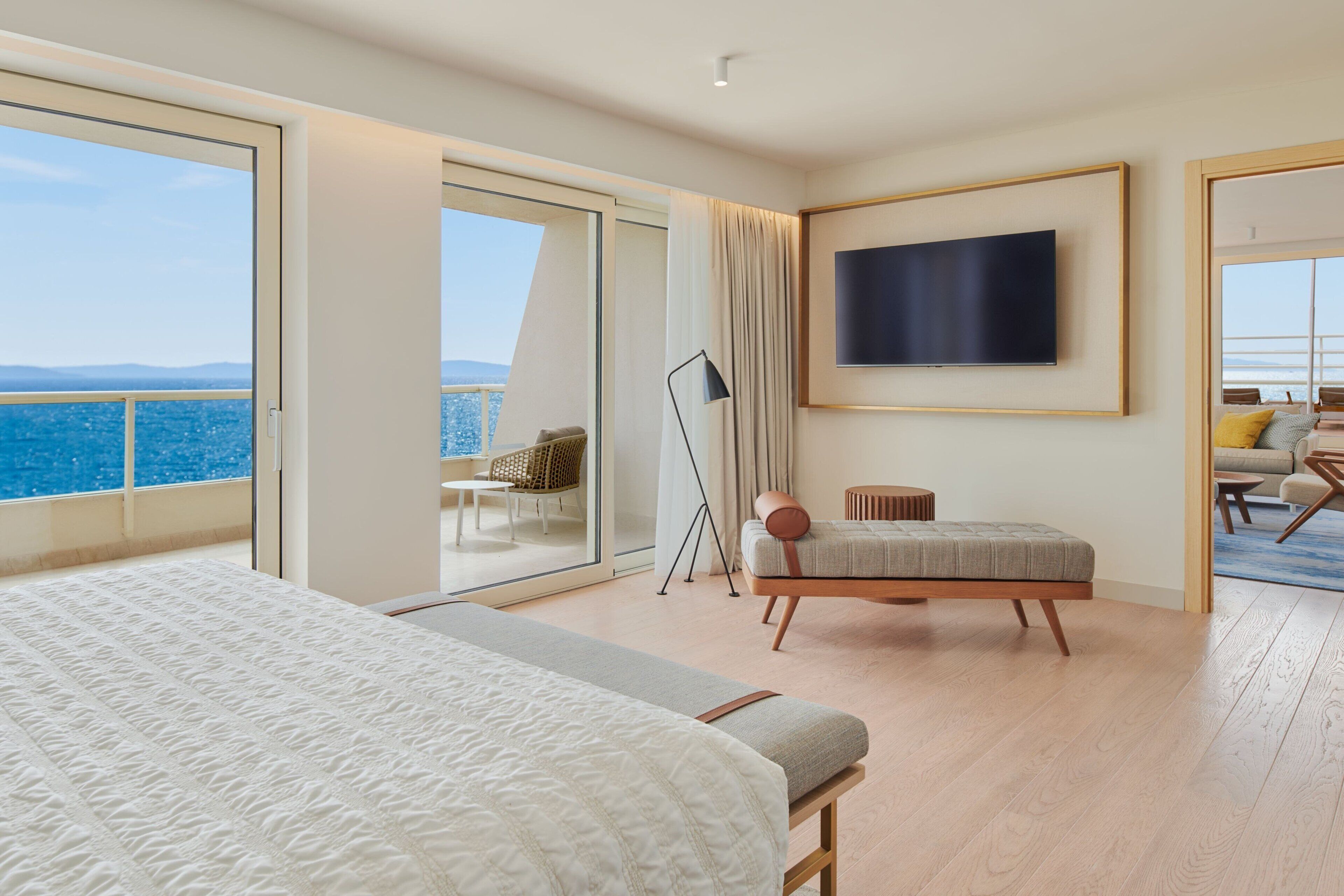 Una habitación de hotel moderna con una cama grande, un espacio de descanso con vistas al mar, un televisor de pantalla plana y un balcón con una silla.