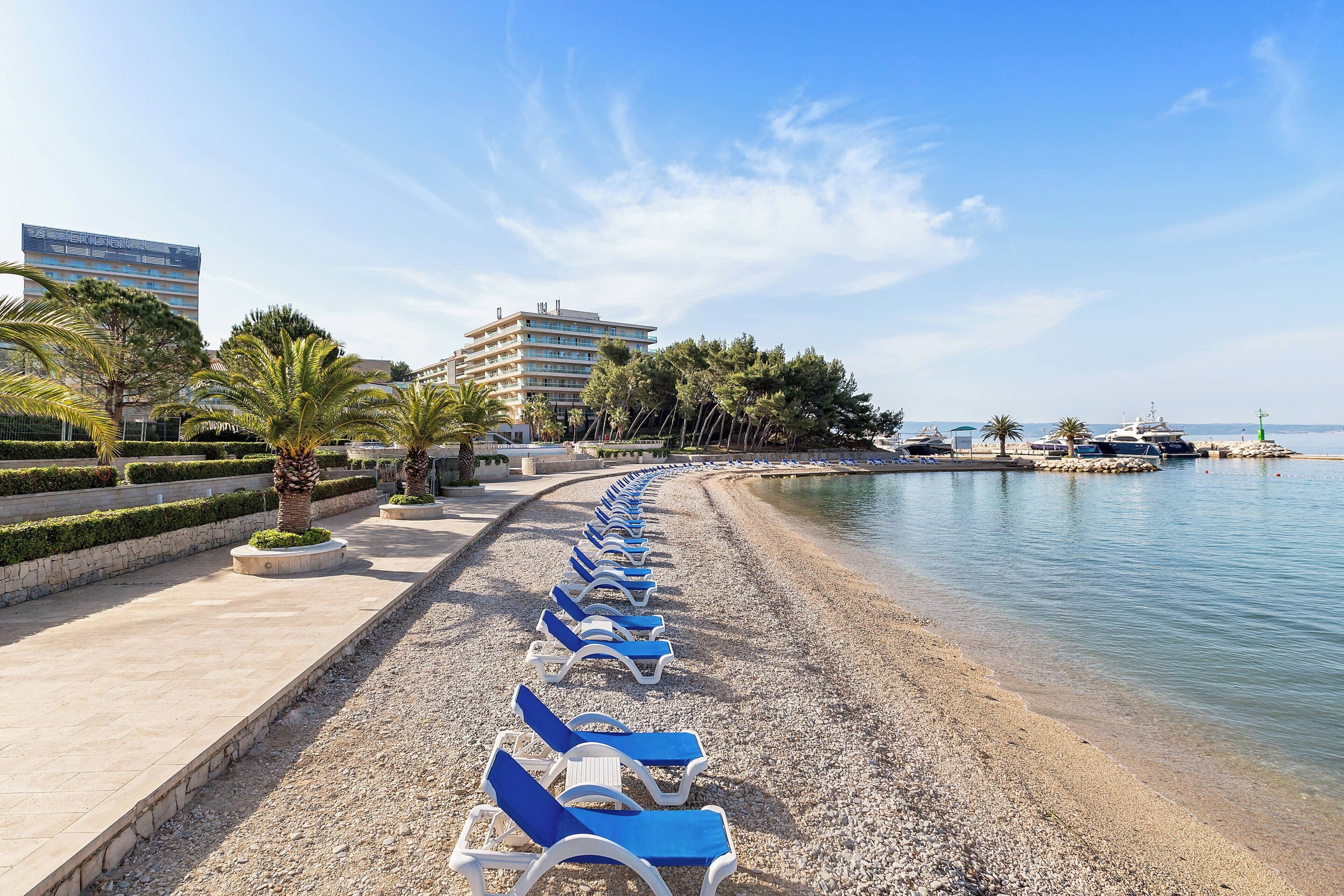 A beachside promenade with rows of sun loungers, a pebble beach, and a calm sea.