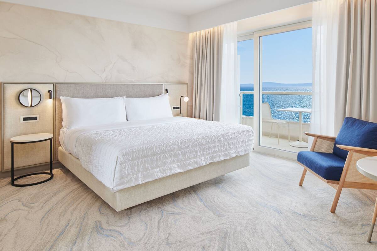 A hotel room with a large bed, a chair, a nightstand, and a view of the sea.