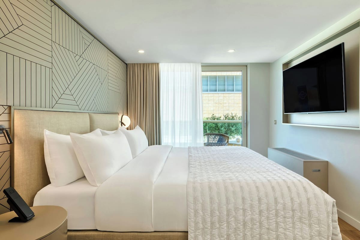 A modern hotel room with a large bed, a flat-screen TV, a bedside table, and a window with a view of greenery.