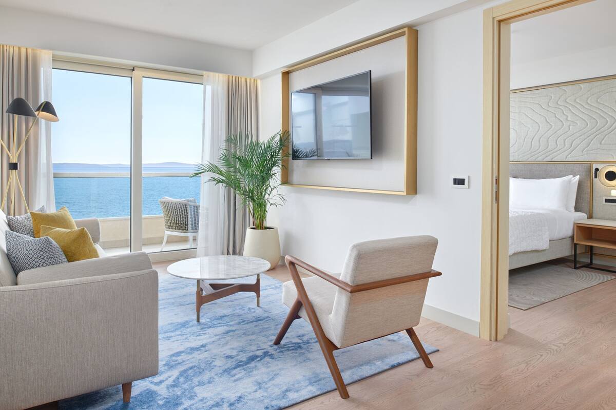 A modern hotel room with a large window offering a view of the sea, a flat-screen TV mounted on the wall, a comfortable sofa, a small round table, and a potted plant.