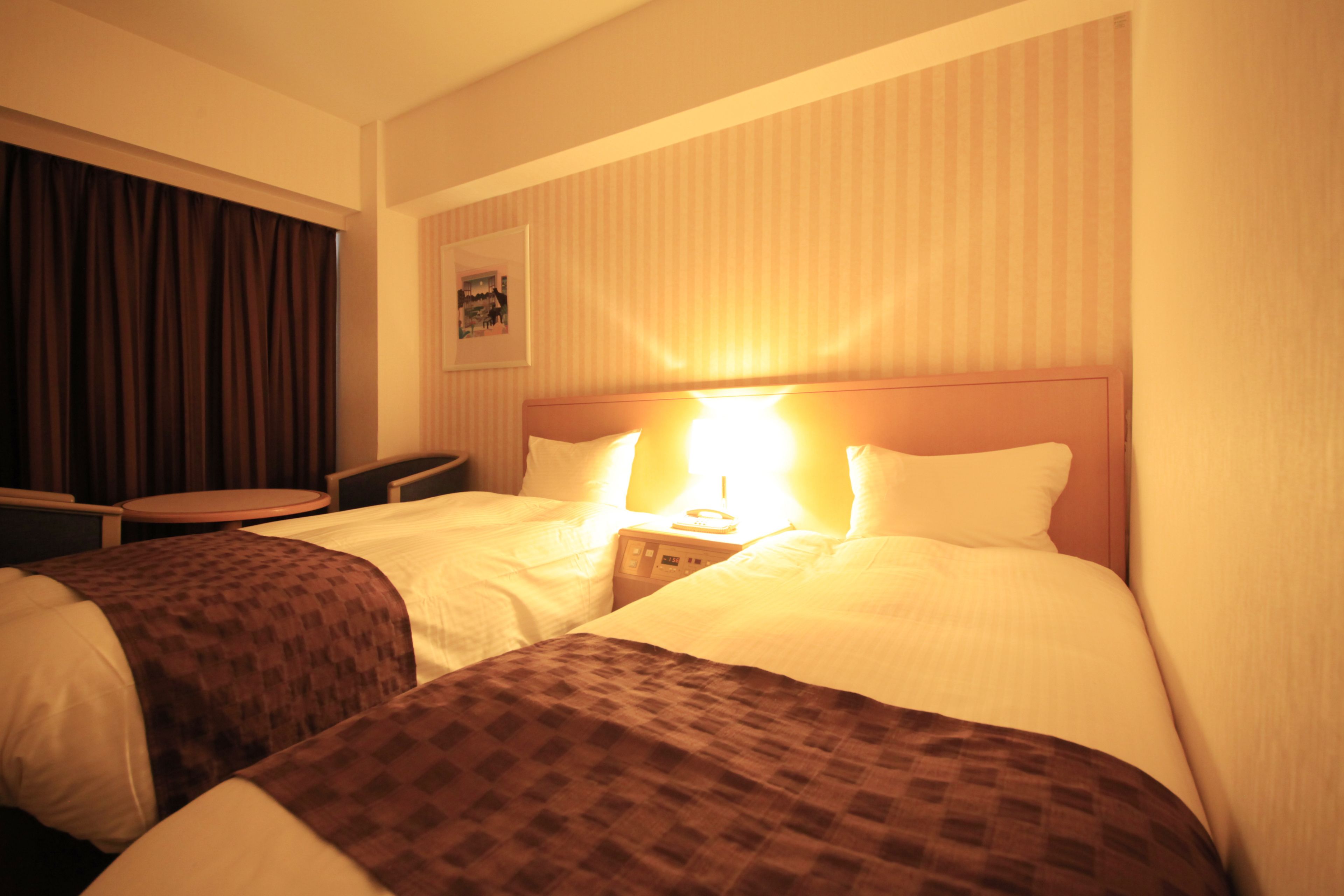 A hotel room with two beds, a bedside table, a lamp, a small table, and a curtain.