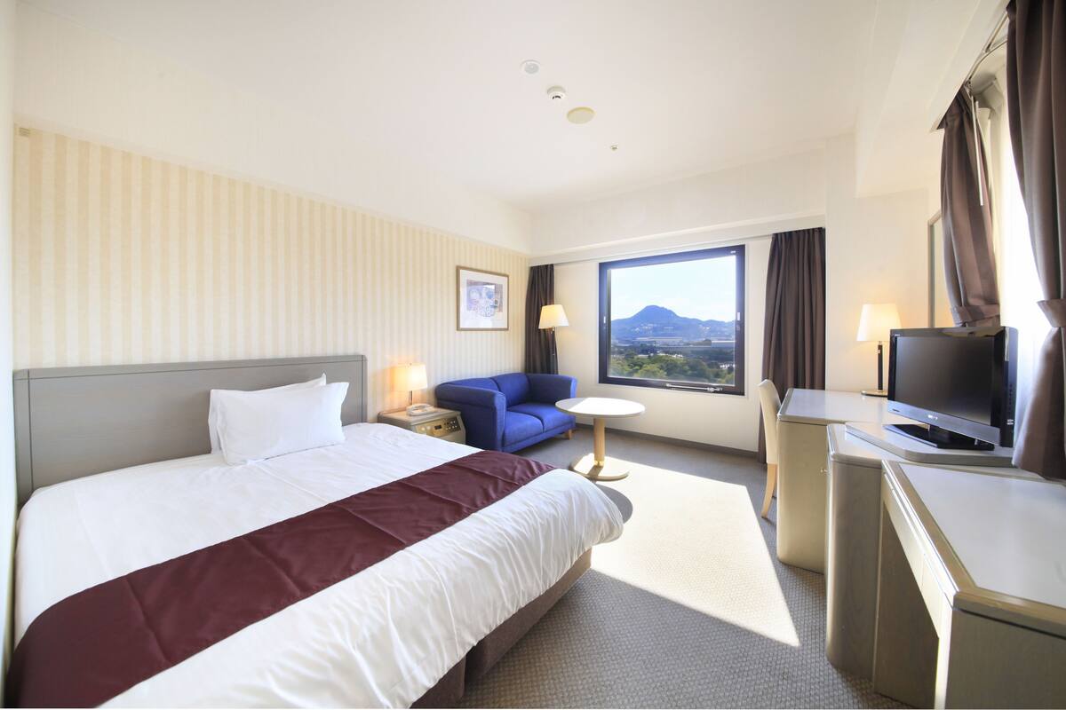 A hotel room with a large bed, a desk with a TV, a small table with a lamp, and a window with a view of mountains.