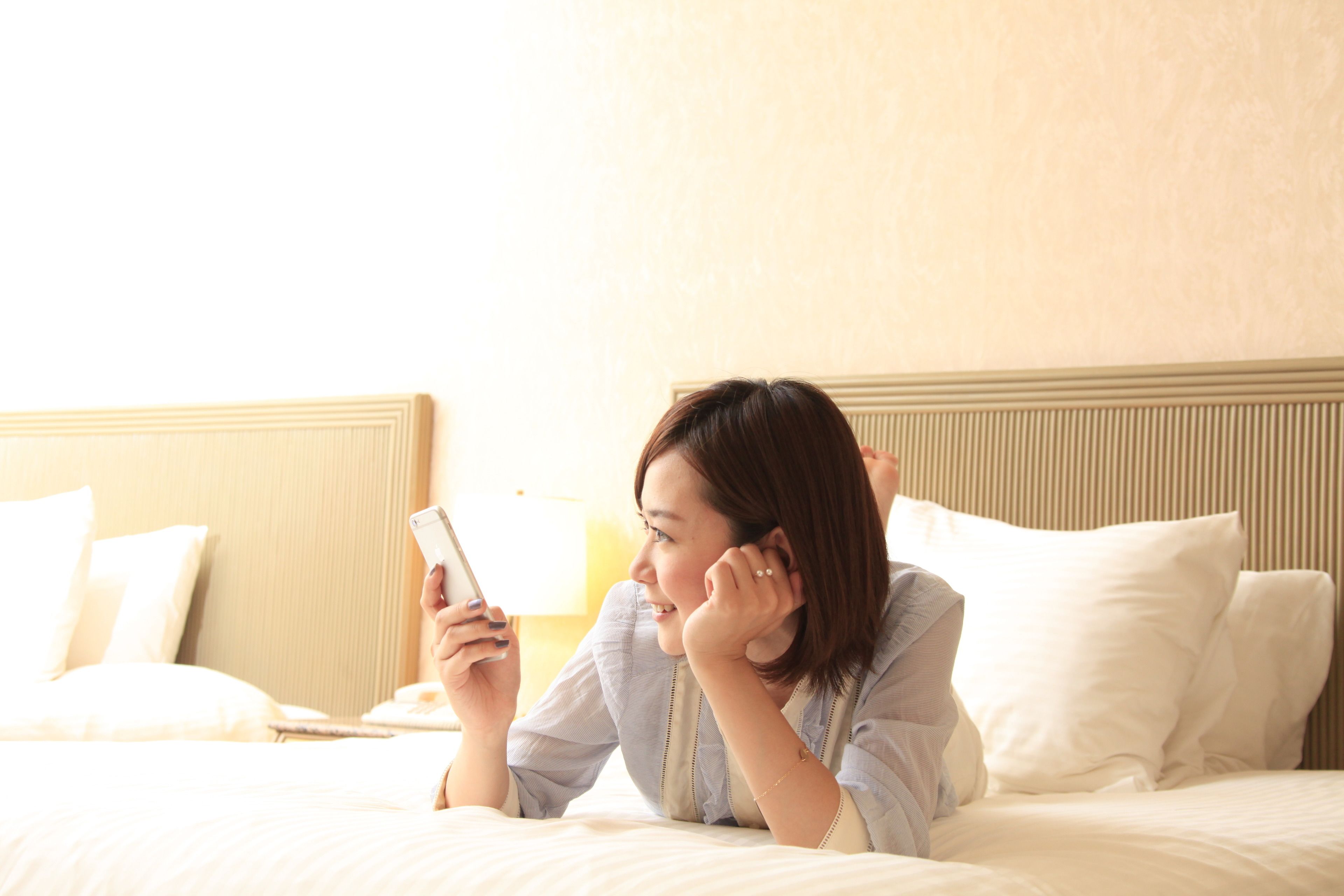 A person lying on a bed in a hotel room, holding a smartphone.