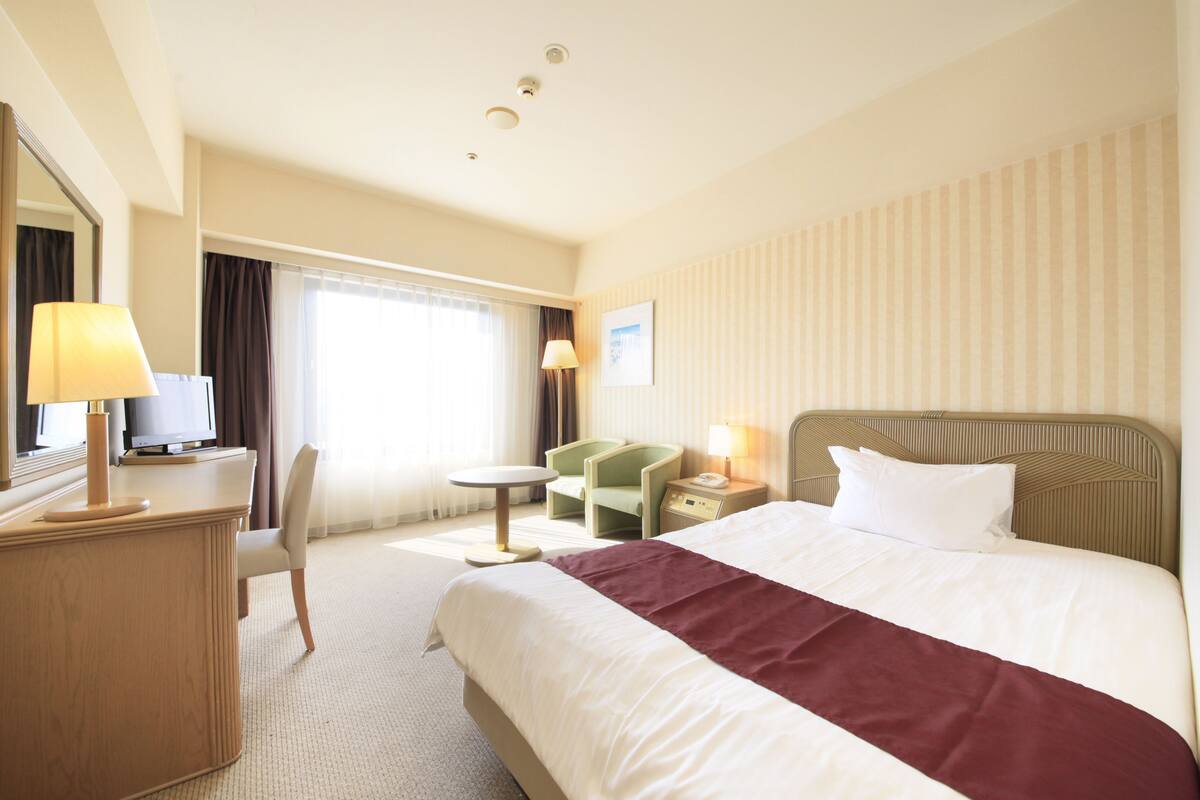 A hotel room with a bed, a desk with a lamp, a chair, a television, and a window with curtains.