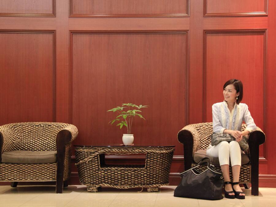 A woman sitting on a patterned armchair in a room with wooden paneled walls and a small table with a plant.