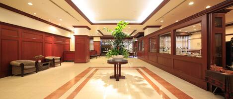 Un hall de hotel con suelo de mármol, paneles de madera, una planta central y zonas de estar.