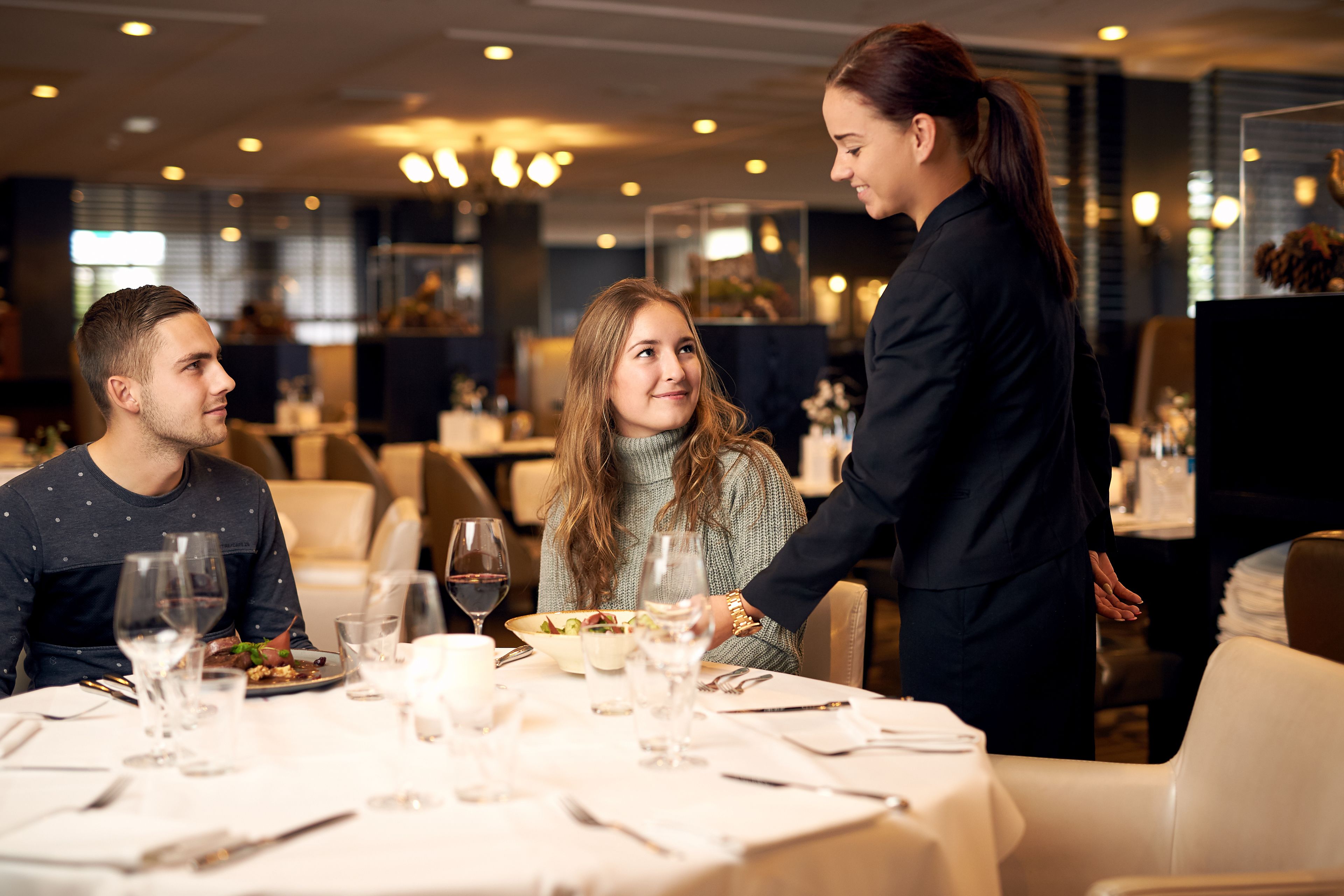 Een eetgedeelte met een tafel voor drie personen, een ober die hen bedient, en een modern interieur met sfeerverlichting.