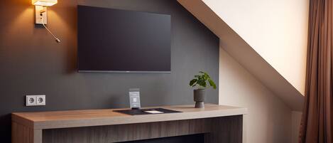 Una habitación de hotel moderna con un televisor de pantalla plana montado en la pared, una mesa consola de madera con una planta y un pequeño otoman.