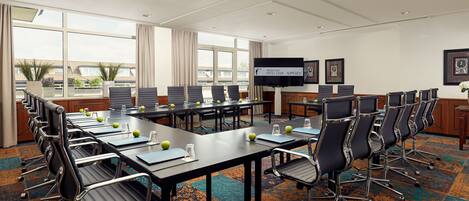 A conference room with multiple tables and chairs, a large screen, and a view of buildings outside.