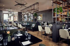 Un restaurante moderno con varias áreas de comedor, mesas y sillas negras. Se observan estanterías con plantas y objetos decorativos.