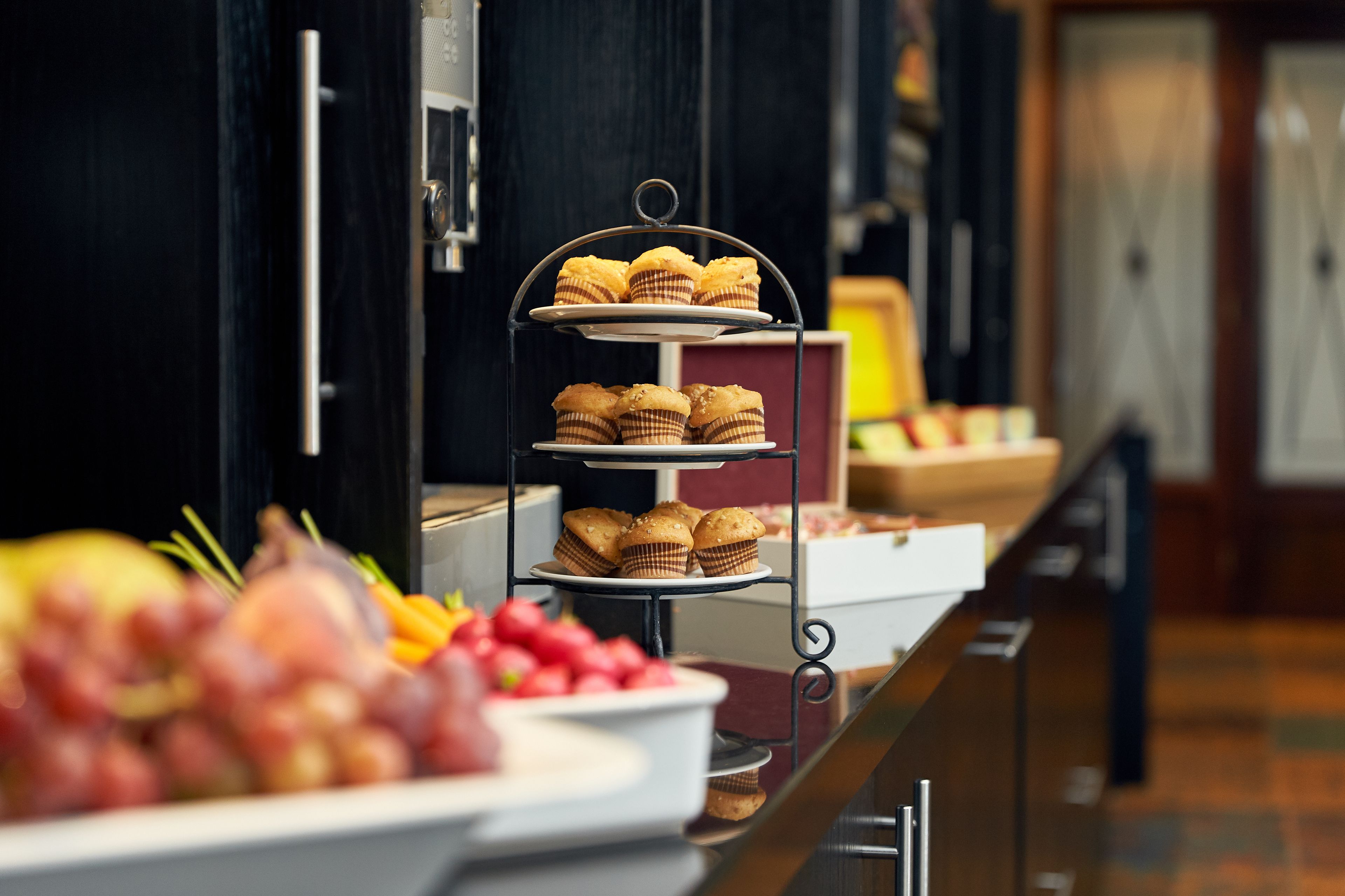 A buffet setup with a three-tiered stand holding muffins and a bowl of fruit in the foreground.