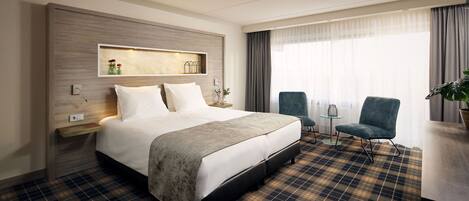 A hotel room with a large bed, a bedside table, a small table with chairs, and a view of the outdoors through a window.
