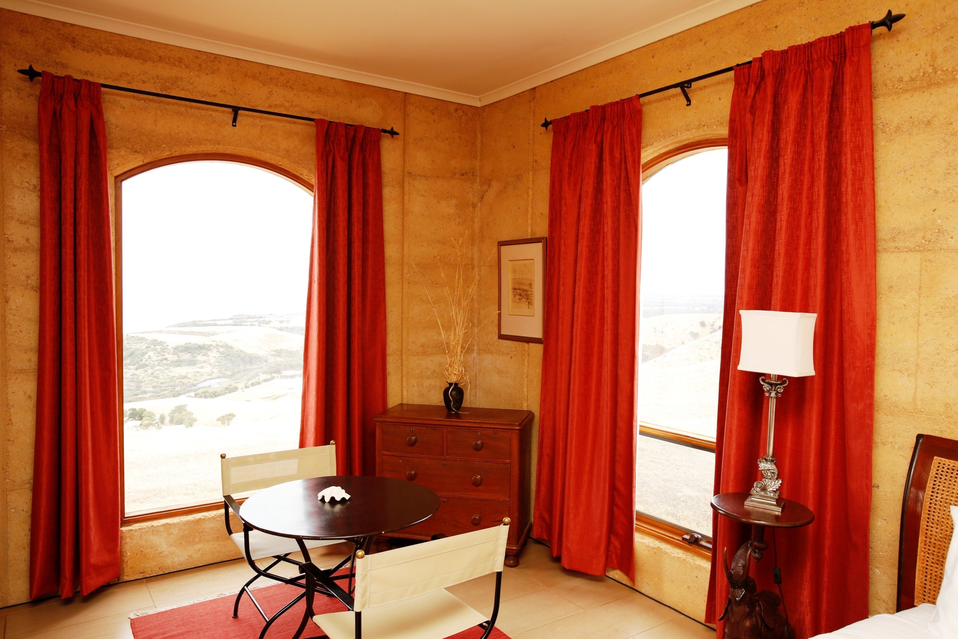 A room with a table and chairs, a dresser, a lamp, and large windows with red curtains.