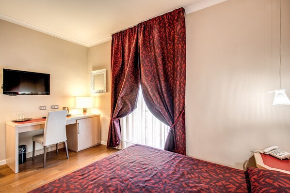 Una habitación de hotel con cama, escritorio, silla, televisor, teléfono y ventana con cortinas.