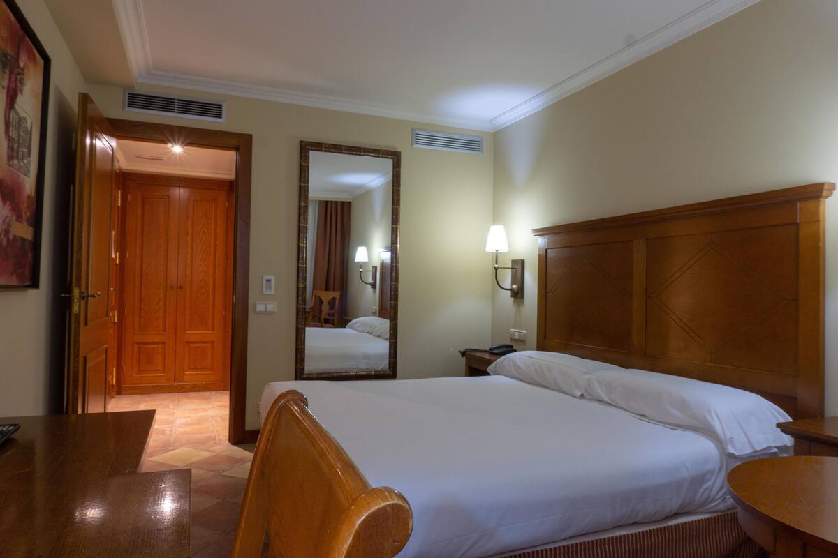 A hotel room with a wooden bed, a nightstand, a mirror, and a door leading to another room.