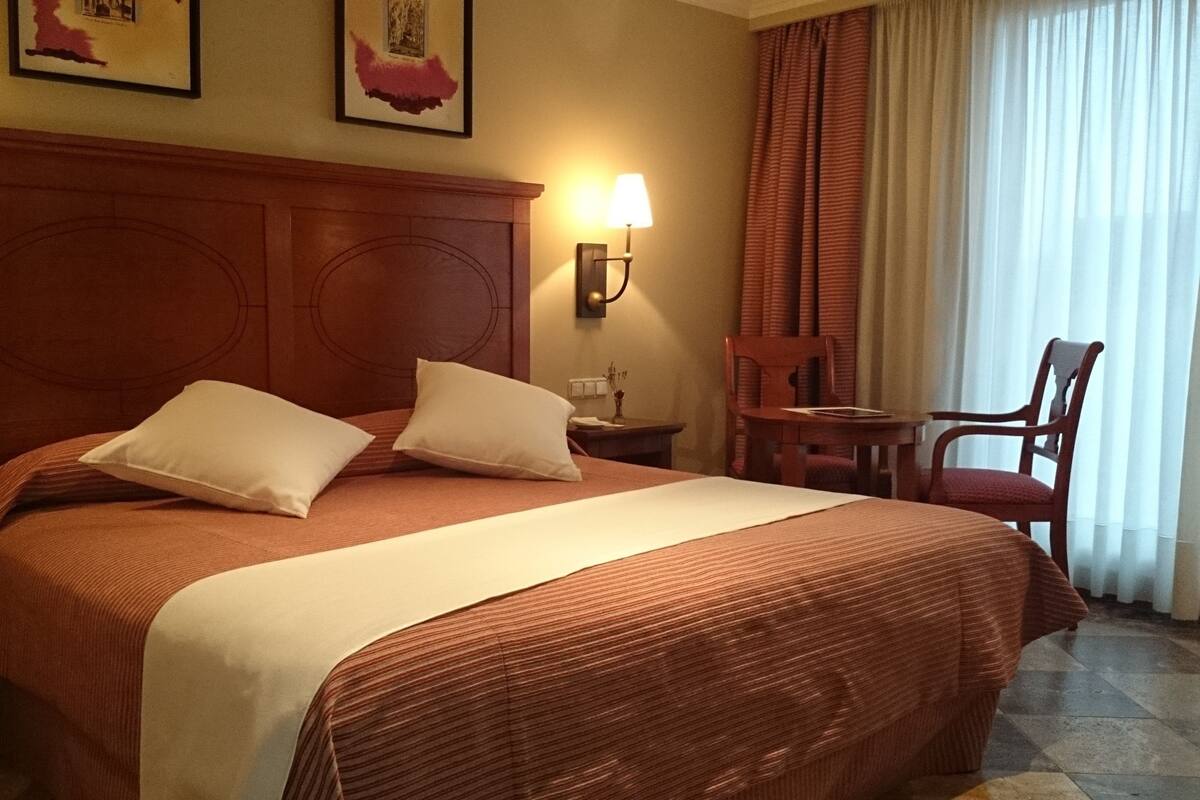 A hotel room with a large bed, two pillows, a nightstand, a lamp, a chair, and a window with curtains.