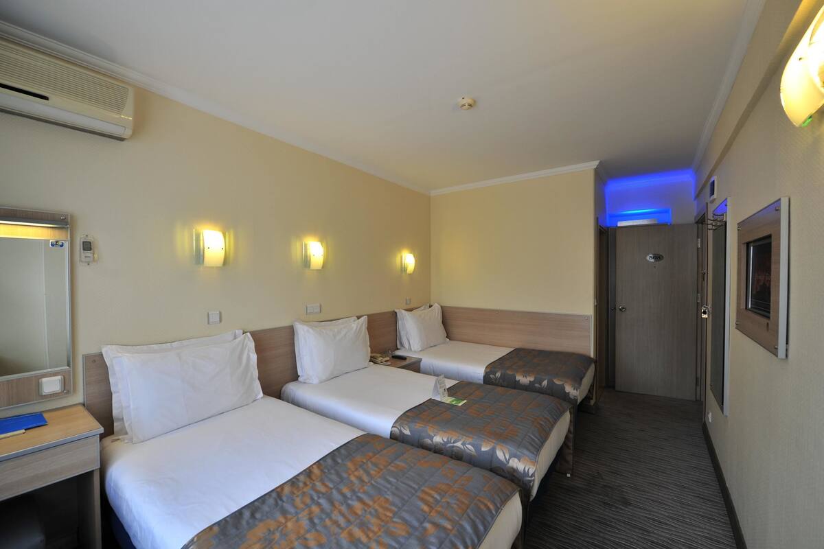 A hotel room with three beds, a large mirror, wall-mounted lights, and a door leading to another room.
