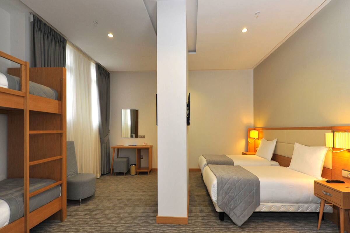 A hotel room with a bunk bed, a desk with a chair, a bedside table with a lamp, and a large bed with pillows and a blanket.