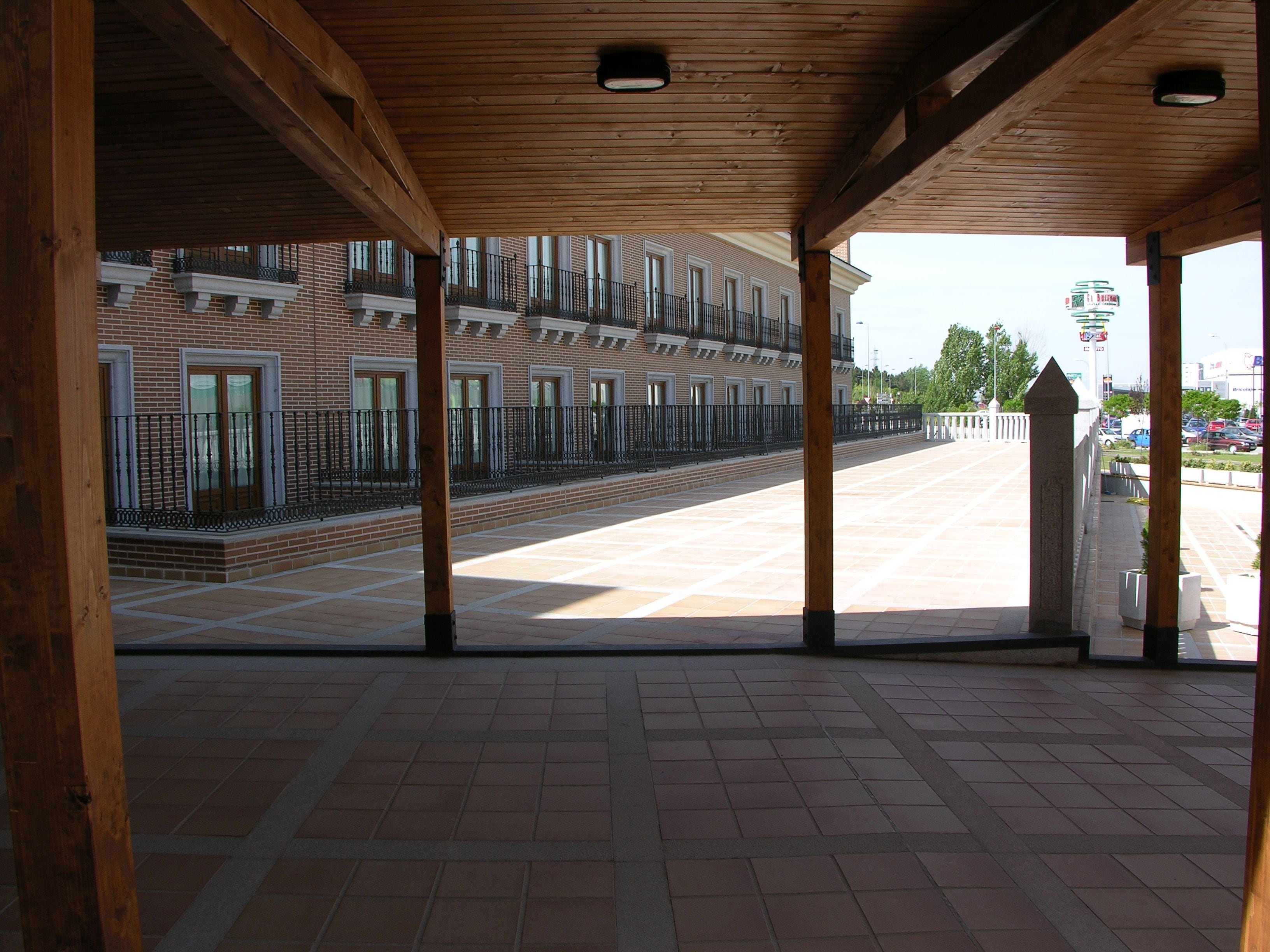 Un espacio descubierto y techado con suelo de baldosas, vigas de madera y vista a un edificio de ladrillo con balcones.
