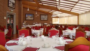 A restaurant with brick walls, wooden beams, and red upholstered chairs. Tables are set with white tablecloths, wine glasses, and cutlery. - Dos Castillas (Ávila)